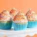 A set of cupcakes with white frosting and orange nonpareil sprinkles in blue wrappers on a white stand.
