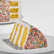 A slice of yellow layer cake with white frosting and covered in colorful rainbow nonpareil sprinkles.