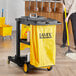 A woman cleaning a floor in a professional kitchen with a Lavex Premium janitor cart with yellow bags and a yellow mop.