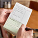 A green and white carbonless guest check pad with beverage lines and a bottom guest receipt, held in a person's hand.