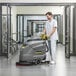 A man in a white uniform using a Karcher walk-behind floor scrubber to clean a floor in a room.