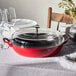 A Staub cherry enameled cast iron pot with a glass lid on a table with wine glasses and a plant.