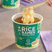 A hand holding chopsticks over a Lotus Foods Tom Yum Rice Ramen Noodle Soup cup.