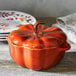 A Staub burnt orange ceramic pumpkin mini Dutch oven on a counter with a stack of plates.