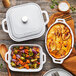 A white Staub baking dish with potatoes and cheese in it on a table with white Staub baking dishes full of vegetables.