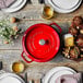 A Staub cherry red enameled cast iron pot on a table with plates and cutlery.