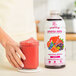 A woman's hand holds a glass of red Smartfruit Superfruit All-Stars puree next to a bottle of the mix.