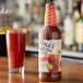 A bottle of Nina's Natural Michelada mix with a white label on a table next to a glass of red liquid.