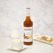 A bottle of Monin brown butter toffee syrup on a white surface next to a white cup and saucer.