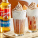 Two glasses of brown drinks with whipped cream on top and a bottle of Torani Pumpkin Pie flavoring syrup.