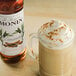 A Monin Premium Cinnamon Syrup bottle next to a glass mug of coffee with whipped cream and cinnamon.