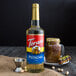 A close up of a Torani Pistachio flavoring syrup bottle on a table with a cup of brown liquid.