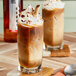 A glass of iced coffee with whipped cream and chocolate on a table with a spoon and a bottle of SHOTT Hazelnut Flavoring Syrup.