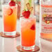 A close-up of two glasses of pink strawberry drinks with a Monin HomeCrafted Strawberry Ginger Lemonade Cocktail mix container on a table.