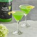 A glass of green Regal Cocktail Apple Martini on a table with a black can of Regal Cocktail Apple Martini mix.