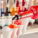 A bottle of red syrup being poured onto a snow cone among several snow cones in paper cups.