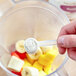 A hand using a measuring spoon to add white Add A Scoop powder to a blender.