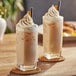Two glasses of Capora Chai Tea Latte with whipped cream and cinnamon on a table.