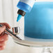 A person using a blue and white Chefmaster airbrush to spray blue food coloring on a cake.