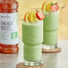 A glass of green smoothie with a lemon and strawberry slice next to a bottle of Monin Energy Boost.