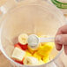 A hand holding a spoon with white Immune Support Blend powder in a blender with fruit.