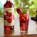 A glass of red liquid with ice and strawberries next to a bottle of Torani strawberry syrup.