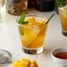 A glass of orange drink with ice, mint leaves, and a straw with a bottle of Monin Premium Honey Mango Flavoring Syrup on the table.