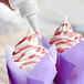 A bottle of purple vanilla glitter glaze being drizzled onto cupcakes with swirled white frosting and purple wrappers.