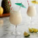 A glass of Monin Pina Colada smoothie with a straw and lime next to another glass with a straw and green umbrella.