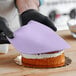 A person wearing black gloves uses a knife to cut a cake covered in Satin Ice lavender vanilla rolled fondant.