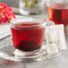 A glass cup of Wild Hibiscus Heart-Tee herbal tea with a tea bag on a saucer.