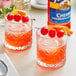 Two glasses of cherry cocktail with cherries and orange slices on a table with a bottle of Torani Sugar-Free Cherry Flavoring Syrup.