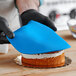 A person wearing black gloves and using a blue spatula to cut a cake covered in blue Satin Ice fondant.