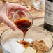 A hand pouring brown liquid from a Hometown Provisions Imitation Vanilla bottle into a glass bowl of brown sugar.