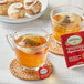 A glass cup of Twinings Gingerbread Joy tea with a tea bag in it, on a coaster with a cinnamon roll.