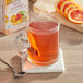 A glass mug of Twinings peach and orange tea with a tea bag in it on a table with a spoon and sliced oranges.