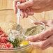 A person holding a spoon of Regal Ground Marjoram over a bowl of meat.