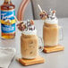 A glass mug filled with brown liquid and topped with whipped cream and chocolate syrup next to cookies.