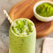 A green smoothie with a straw and a bowl of green matcha powder on a table.