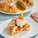 A slice of apple pie with whipped cream being drizzled with caramel sauce on a white plate.