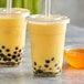 Two cups of yellow bubble tea with black balls on a table with a bowl of orange liquid.