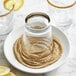 A glass jar with a brown rim and a lid filled with Regal Celery Salt with a lemon slice on a plate of sand.