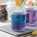 A glass mug of blue butterfly pea flower tea with lemon being squeezed into it, next to another mug and a jar of dried tea flowers.
