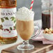A glass of brown liquid with whipped cream and a red and white straw next to a bottle of Monin Hazelnut Syrup.
