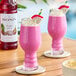 A bottle of Monin Premium Dragon Fruit Flavoring Syrup next to two glasses of pink dragon fruit smoothies garnished with whipped cream and dragon fruit slices.