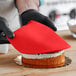 A person wearing black gloves and using a red spatula to cut a cake covered in red Satin Ice fondant.