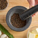 A stone mortar and pestle filled with whole black peppercorns on a wooden surface, surrounded by herbs, garlic, and butter.