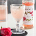 A wine glass of pink liquid with a Monin Rose Flavoring Syrup bottle on a table next to a rose.