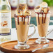 Two glasses of hot chocolate with marshmallows and Capora Toasted Marshmallow flavoring syrup on a table in a coffee shop.