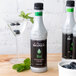 A white Monin Mint Concentrated Flavor bottle with a black label on a table next to a glass of blueberries.